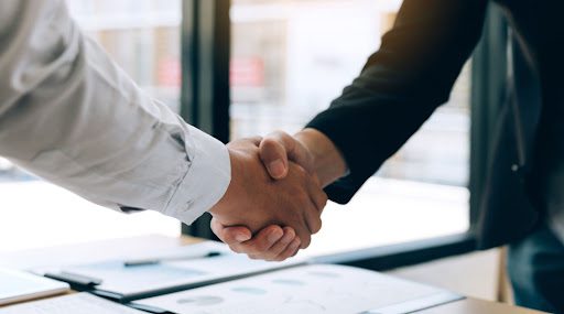 two business owners shake hands over a desk in an office.